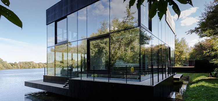 Pasakaina stikla māja uz ezera, kurā atspoguļojas apkārtnes ainava